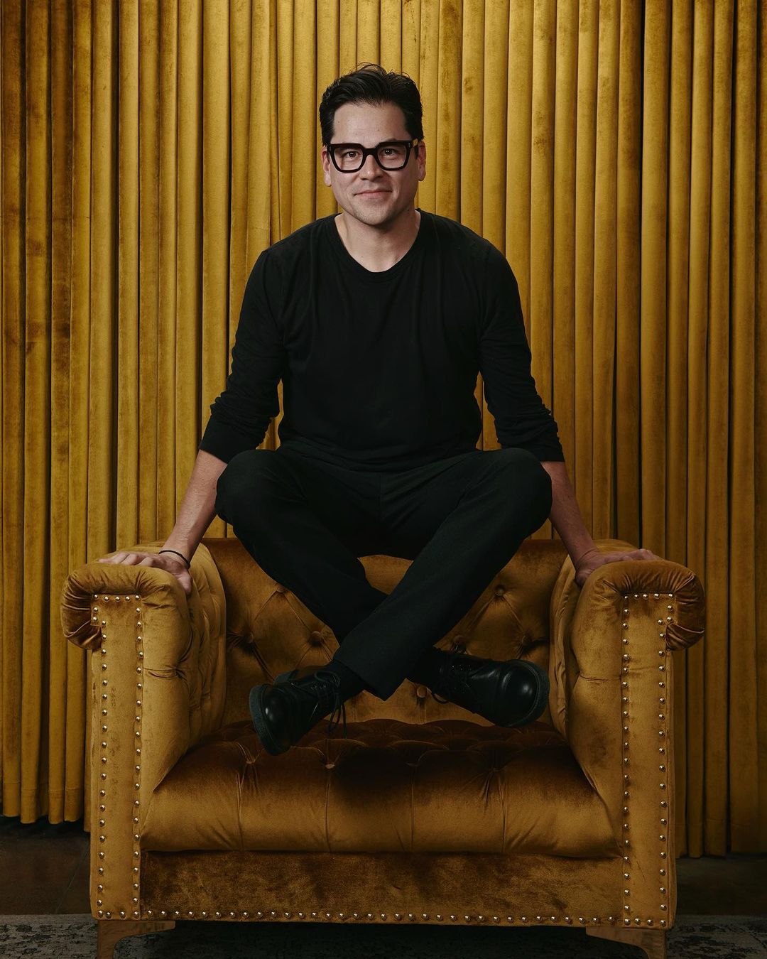 An adult male with dark hair, wearing glasses and a black outfit, sits cross-legged in a yellow velvet tufted armchair in an indoor studio setting.